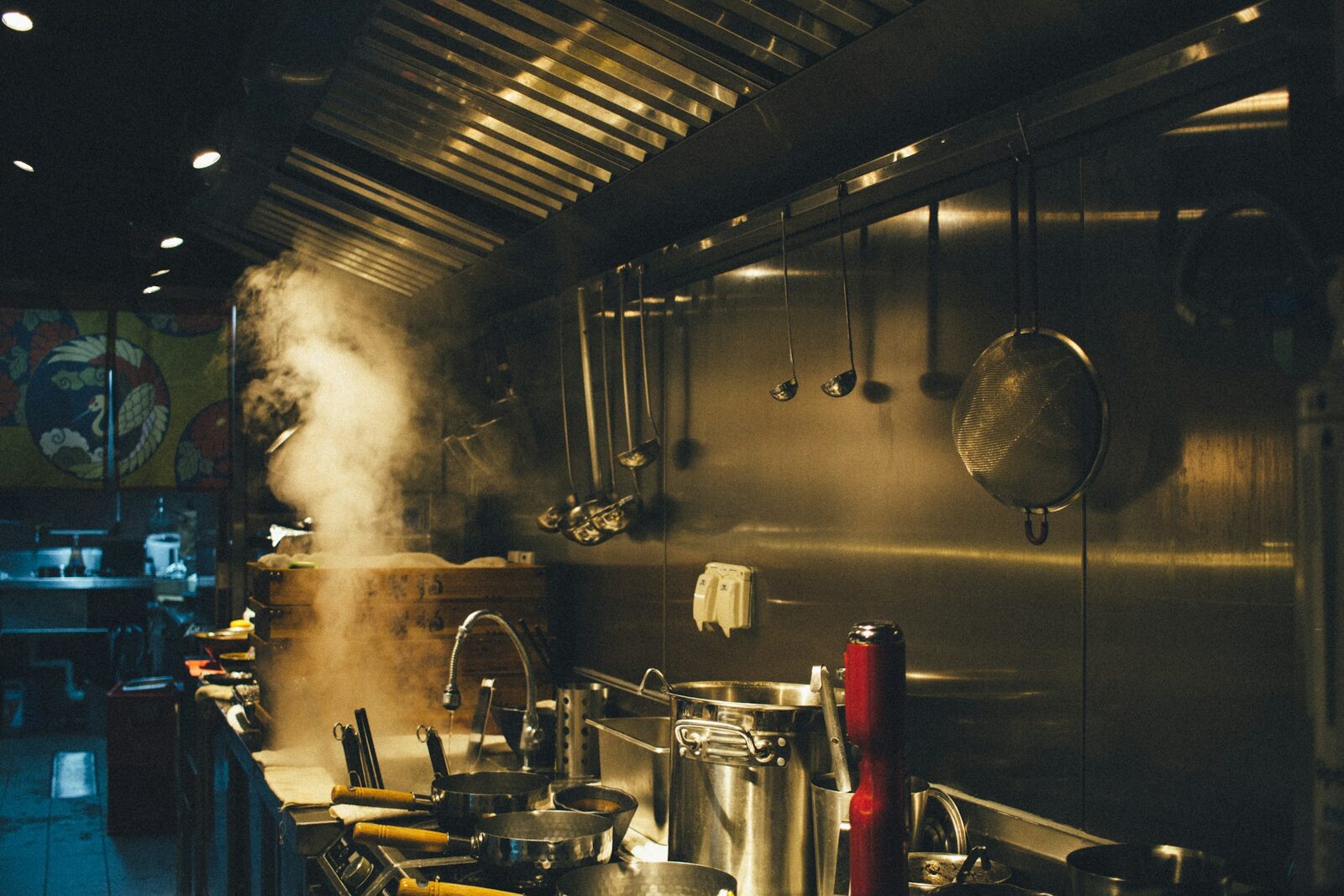 A stainless steel commercial kitchen line with an extractor hood, hanging utensils and steaming pots